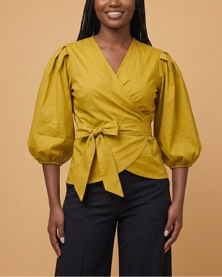 A woman modeling an olive colored wrap-style blouse with puffed sleeves, paired with black pants, against a solid beige background.