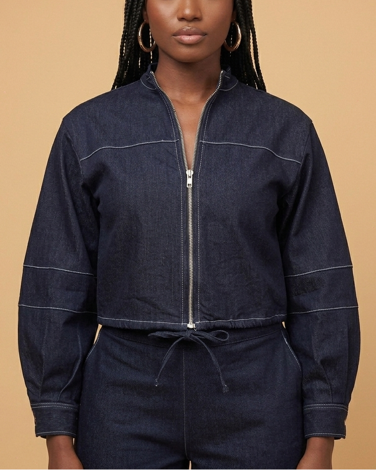A woman models a dark navy denim jacket with a silver zipper, contrast white stitching, and a drawstring waist, set against a solid beige background.