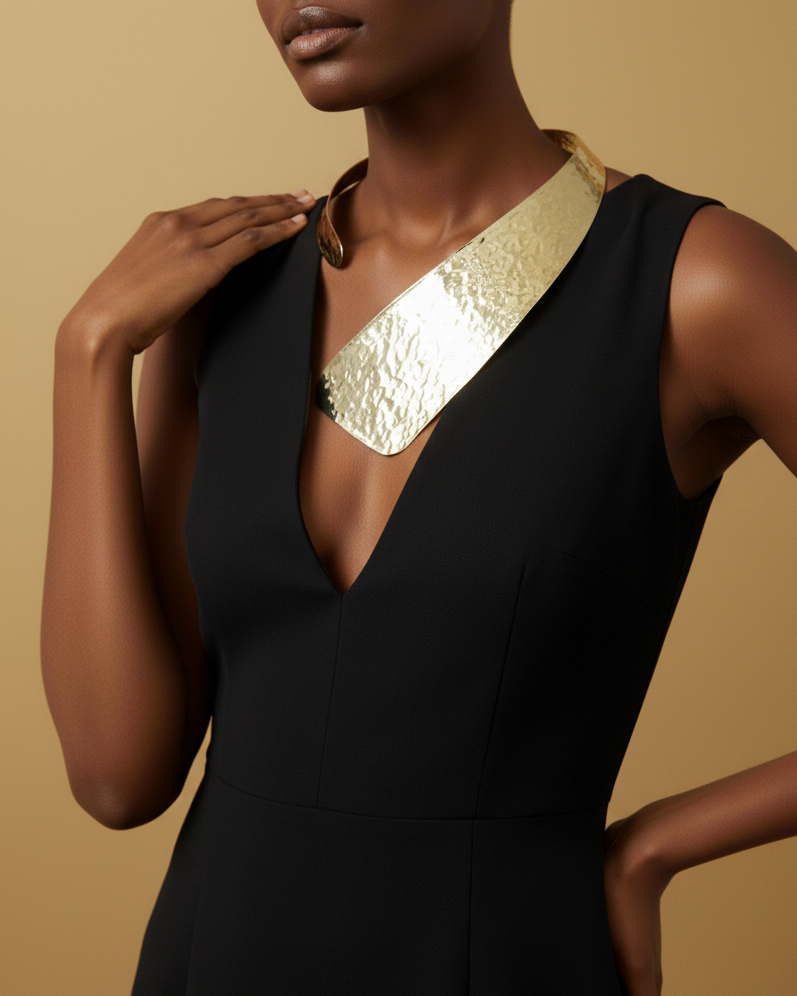 Model wearing a black dress with a gold statement neckpiece against a beige background. 